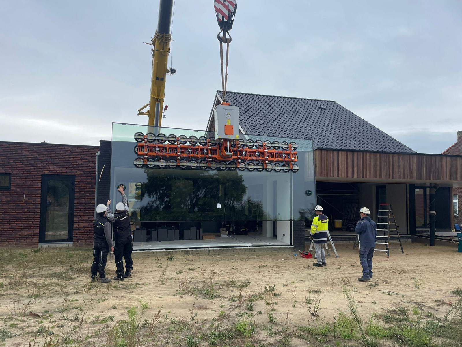 Hijskraan tilt een grote glasplaat in de gevel van een woning in aanbouw.